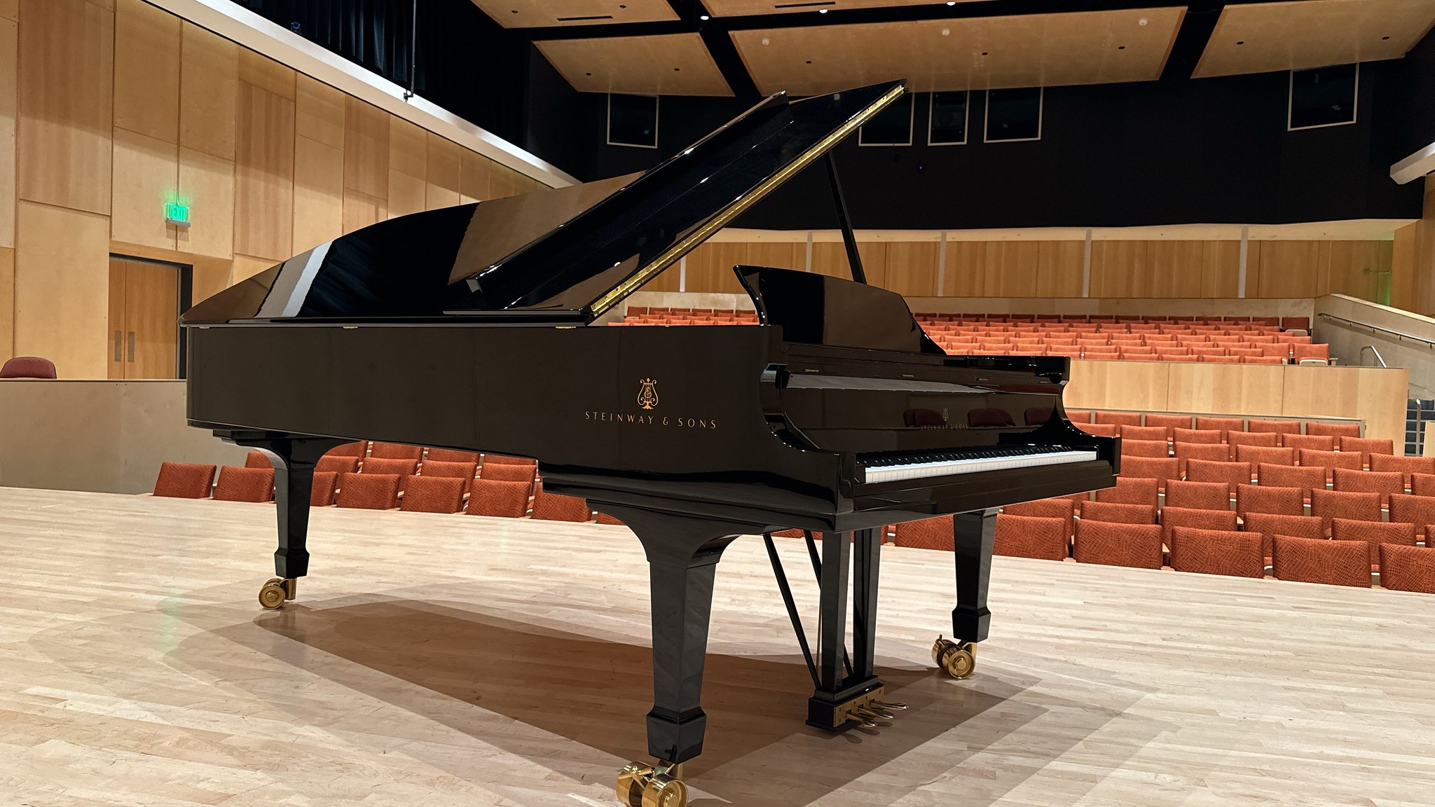 Black Steinway & Sons grand piano on the stage of Boyd Concert Hall at Allan Hancock College’s Santa Maria campus. The piano sits on a wooden stage with its lid open, facing rows of empty orange auditorium seats in a modern concert hall setting.